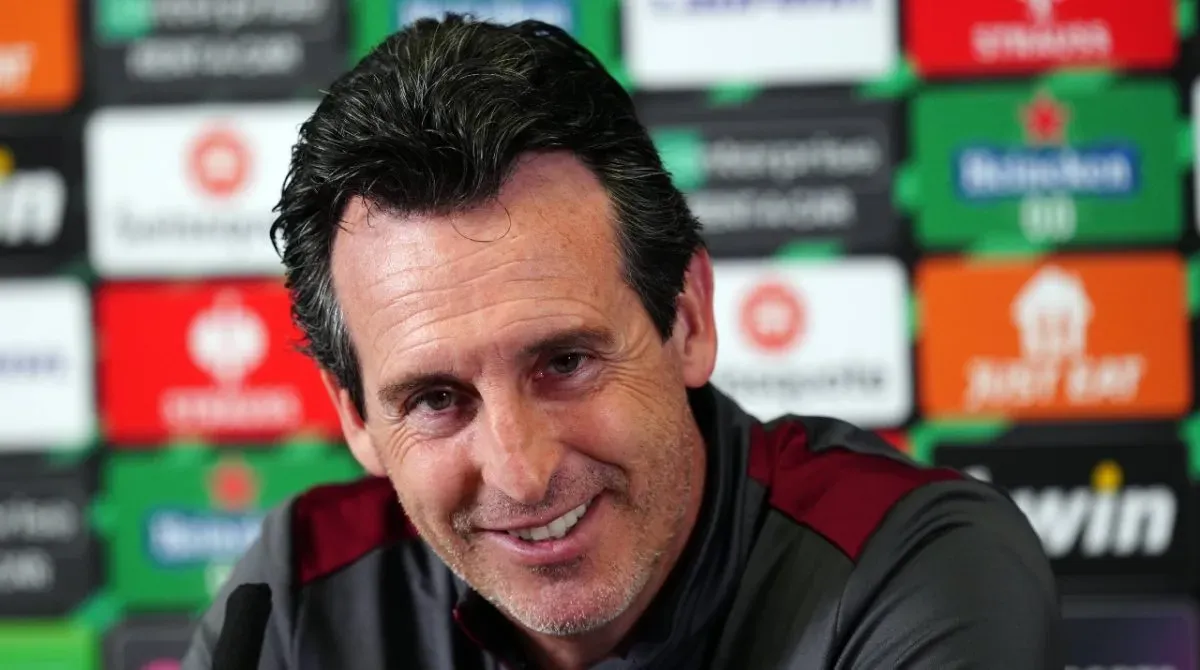 A smiling Unai Emery sits in front of a board of Conference League sponsors.